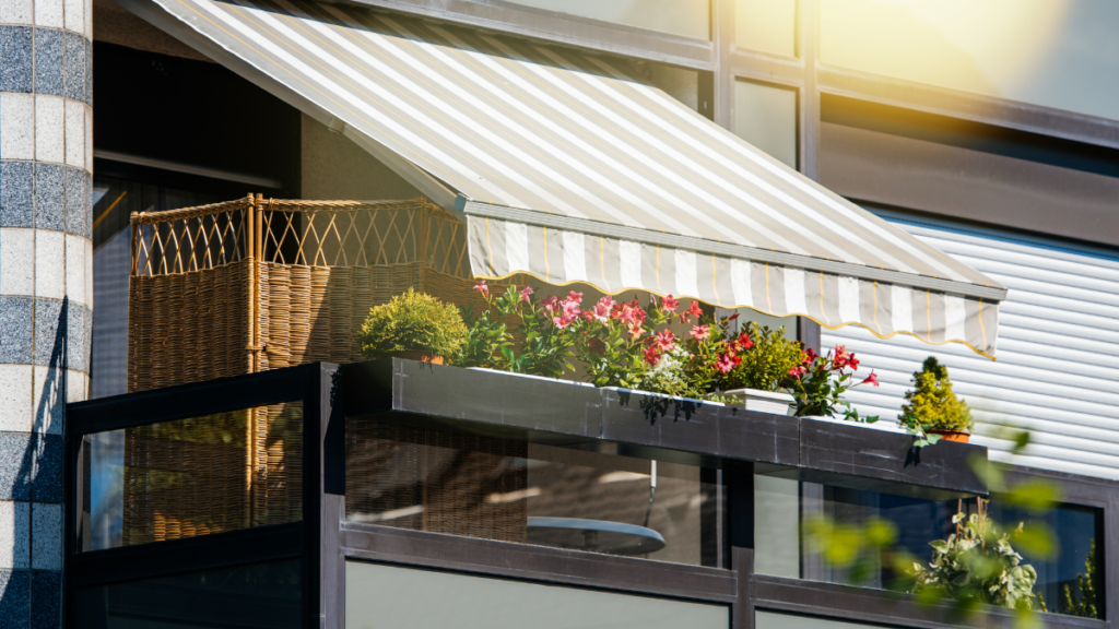 Mały balkon w bloku z markizą, skrzynkami z kwiatami oraz osłoną z rattanu